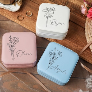 Peut inclure: Trois bo&icirc;tes &agrave; bijoux carr&eacute;es blanches, roses et bleues. Chaque bo&icirc;te pr&eacute;sente un motif floral et un nom en &eacute;criture cursive : Regina, Olivia et Angela. Les bo&icirc;tes sont zipp&eacute;es et pos&eacute;es sur une surface en bois.