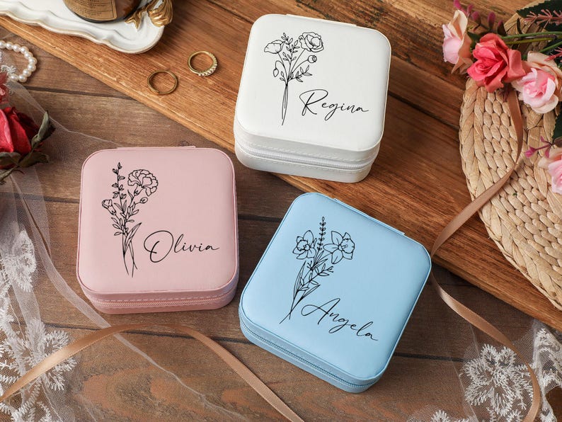 Peut inclure: Trois bo&icirc;tes &agrave; bijoux carr&eacute;es blanches, roses et bleues. Chaque bo&icirc;te pr&eacute;sente un motif floral et un nom en &eacute;criture cursive : Regina, Olivia et Angela. Les bo&icirc;tes sont zipp&eacute;es et pos&eacute;es sur une surface en bois.