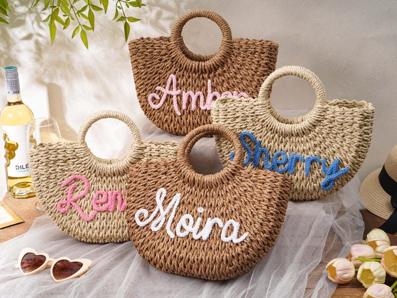 May include: Four woven straw handbags with round handles, each featuring a personalized name embroidered in cursive yarn. The names Amber, Sherry, Ren, and Moira are displayed in pink, blue, and white colors on the front of the textured bags.