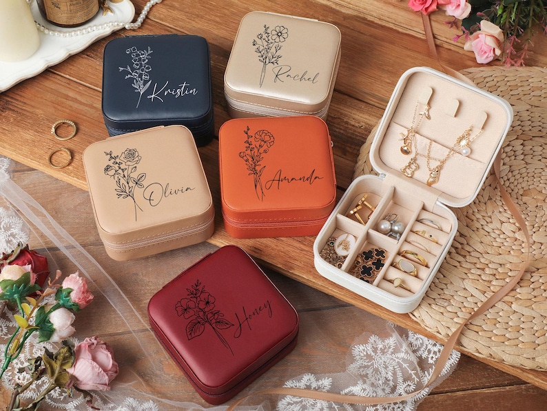 Peut inclure: Plusieurs bo&icirc;tes &agrave; bijoux carr&eacute;es personnalis&eacute;es, de couleurs vari&eacute;es : bleu marine, beige, orange et bordeaux. Chaque bo&icirc;te est orn&eacute;e d'un motif floral et d'un nom. Une bo&icirc;te est ouverte, r&eacute;v&eacute;lant des compartiments.