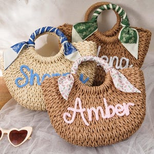 May include: Three woven straw handbags with rounded handles wrapped in patterned fabric scarves. Each bag features a name embroidered in cursive yarn: one in light blue reading "Sherry", one in white reading "Elena", and one in pink reading "Amber".