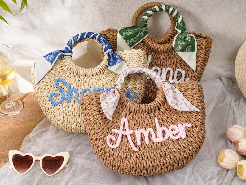 May include: Three woven straw handbags with rounded handles wrapped in patterned fabric scarves. Each bag features a name embroidered in cursive yarn: one in light blue reading "Sherry", one in white reading "Elena", and one in pink reading "Amber".
