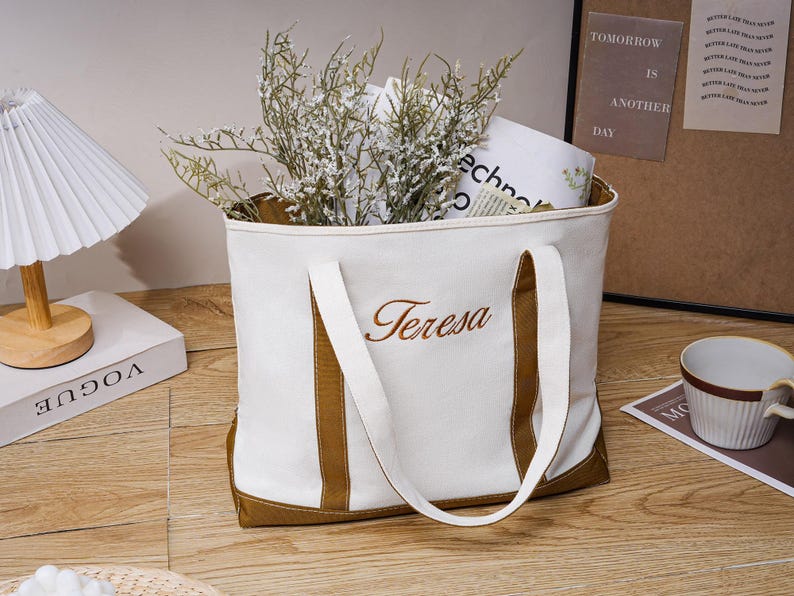 以下が含まれることがあります： 茶色のアクセントと「Teresa」の刺繍が入った白いキャンバスのトートバッグ。バッグには緑と本が入っています。左側には白いランプと「Vogue」の本があります。