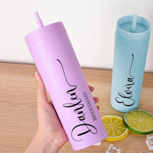 May include: Two personalized tumblers with straws. One is lavender with the name "Parker" and the word "Bridesmaid" in black script. The other is light blue with the name "Elvira" in black script. Decorative fruit slices and ice cubes are also visible.