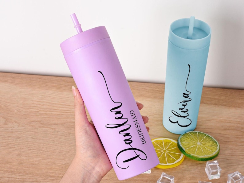 May include: Two personalized tumblers with straws. One is lavender with the name "Parker" and the word "Bridesmaid" in black script. The other is light blue with the name "Elvira" in black script. Decorative fruit slices and ice cubes are also visible.