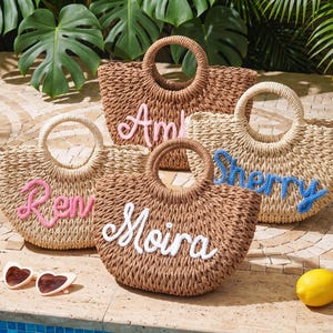 May include: Four woven straw tote bags with round handles, each featuring a personalized name in cursive script. The bags are in shades of tan and brown, with names embroidered in pink, blue, and white yarn. Heart-shaped sunglasses and lemons are nearby.