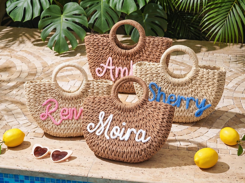 May include: Four woven straw tote bags with round handles, each featuring a personalized name in cursive script. The bags are in shades of tan and brown, with names embroidered in pink, blue, and white yarn. Heart-shaped sunglasses and lemons are nearby.