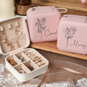 Może przedstawiać: Otwarta biała szkatułka na biżuterię prezentuje różne złote kolczyki, naszyjniki i pierścionki. Widoczne są również dwa różowe etui na biżuterię z kwiatowymi wzorami i napisami "Coral" i "Meroy". Etui znajdują się na drewnianej powierzchni.