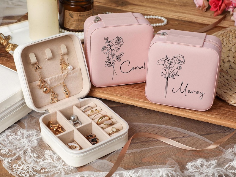 Peut inclure: Une bo&icirc;te &agrave; bijoux blanche ouverte pr&eacute;sente des boucles d'oreilles, colliers et bagues dor&eacute;s. Deux bo&icirc;tes &agrave; bijoux roses avec des motifs floraux et les noms "Coral" et "Meroy" sont &eacute;galement visibles. Les bo&icirc;tes sont sur une surface en bois.