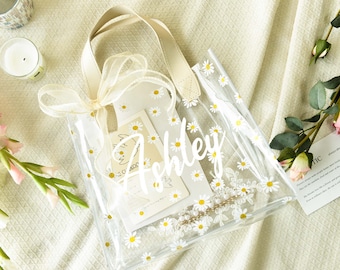 May include: Clear plastic tote bag with a white daisy print and the name "Ashley" written in white script. The bag has a beige handle and a white ribbon tied around the handle.