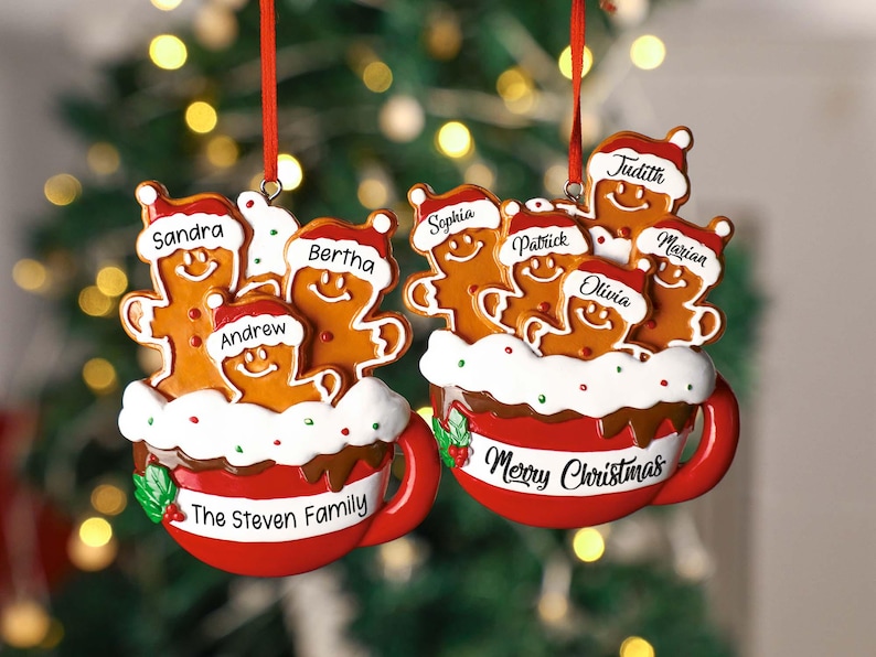 May include: Two personalized Christmas ornaments feature gingerbread families in red mugs.  One mug says 'The Steven Family' and the other says 'Merry Christmas.' Gingerbread figures are decorated with names like Sandra, Andrew, Bertha, Sophia, Patrick, Olivia, Judith, and Marian.  The ornaments are red, white, and brown, with green holly accents.