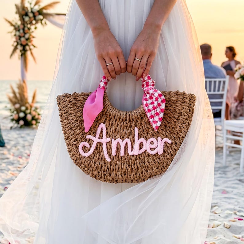 May include: A crescent-shaped woven straw handbag featuring the name Amber in light pink script lettering. The handles are adorned with two fabric ribbons, one solid pink and one pink and white gingham check, tied in a decorative knot style.