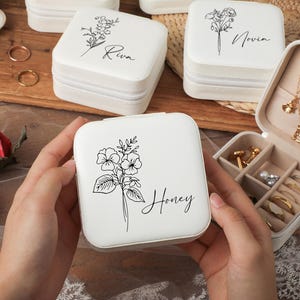 Peut inclure: Bo&icirc;tes &agrave; bijoux carr&eacute;es blanches avec des motifs floraux et des noms personnalis&eacute;s. Une bo&icirc;te est ouverte, r&eacute;v&eacute;lant des compartiments pour bagues, boucles d'oreilles et colliers. D'autres bo&icirc;tes sont ferm&eacute;es, avec des noms comme "Honey" et "Riva".