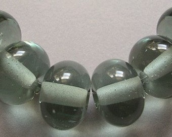May include: A strand of translucent, gray-green glass beads. Each bead has a rounded shape with a lighter, opaque band encircling the center. The beads are strung on a thin, white cord, suitable for jewelry making.