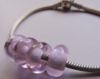 May include: A silver bracelet featuring four translucent, lilac-coloured glass beads. The beads are round and have a slightly frosted appearance, threaded onto a silver chain. The bracelet is set against a soft, neutral background.