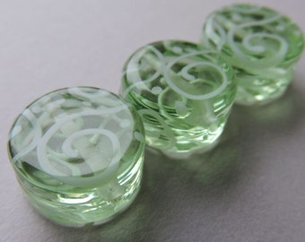 May include: Three round, light green glass objects with white, swirling designs. The objects are arranged in a row, with the closest one in focus. The glass has a slightly faceted appearance, and the white design is etched into the surface.