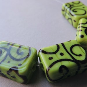 May include: Lime green square beads with dark brown swirling patterns. The beads are strung together on a thin, clear string. The beads have rounded corners and a glossy finish, ideal for jewellery making.