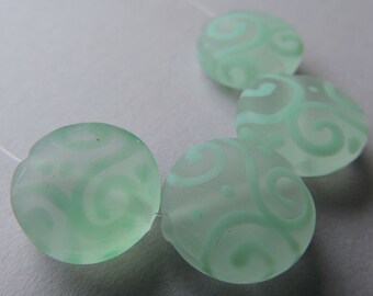 May include: Four frosted, light green glass beads with a flat, oval shape. Each bead features a swirling, white pattern. The beads are strung on a thin, white thread, suggesting they are for jewelry making or crafts.