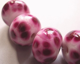 May include: A string of four round glass beads featuring a glossy white base with irregular, deep magenta and dark purple spots. Each bead has a central hole for threading, and the surface of the glass reflects light, showing a smooth, polished finish.