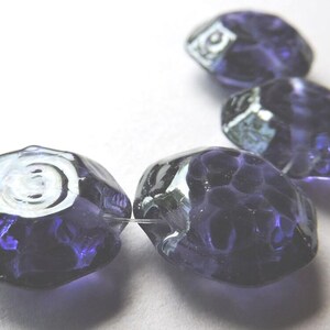 May include: Close-up of four faceted, dark purple beads. The beads have an irregular shape and a textured surface. The beads are strung on a clear thread, and are set against a white background. These beads are suitable for jewelry making.