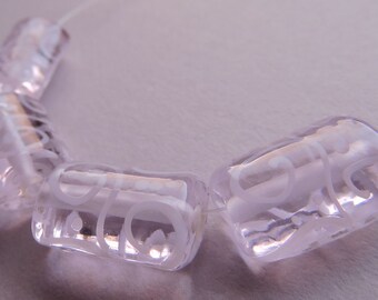 May include: Close-up of a strand of clear, rectangular beads with etched white designs. The beads are strung on a thin, clear cord. The beads have a soft, translucent appearance, and the etched designs add a decorative element.