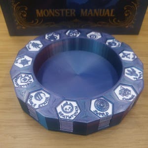 Peut inclure: Un plateau bleu et violet imprimé en 3D avec un design circulaire. Le plateau présente une série de carreaux hexagonaux blancs avec divers symboles et du texte. Le texte "MONSTER MANUAL" est visible en arrière-plan.