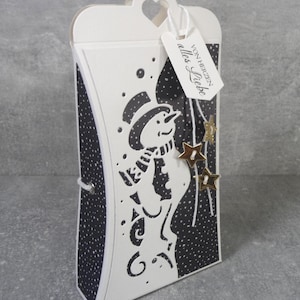 Könnte beinhalten: Eine weiße kissenförmige Geschenkbox mit einer schwarzen, ausgestanzten Schneemann-Silhouette auf schwarzem Hintergrund mit weißen Punkten. Die Box hat eine herzförmige Ausstanzung oben und ein weißes Etikett mit dem Text "VON HERZEN alles Liebe".