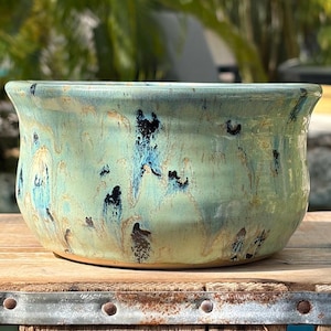 May include: A round ceramic bowl featuring a glossy glaze in shades of light green, turquoise, and dark blue. The surface displays a marbled, drip-like pattern with visible streaks of tan and brown throughout the exterior of the vessel.