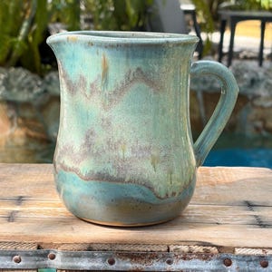 May include: A ceramic pitcher featuring a glossy glaze in shades of turquoise, seafoam green, and muted brown. The vessel has a wide, rounded body, a curved handle on the right side, and a small pouring spout at the top rim with a smooth finish.