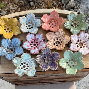 May include: A collection of eleven ceramic flower-shaped soap dishes in various colors including yellow, blue, pink, green, and purple. Each dish features a five-petal design with a central cluster of drainage holes and a glossy glazed finish.