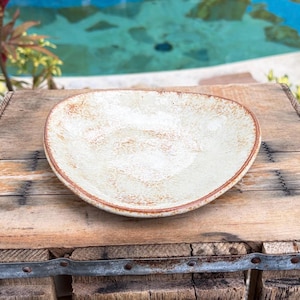 May include: A cream-coloured, irregularly shaped ceramic plate with a brown rim. The plate has a textured surface and is set on a rustic wooden surface. The plate is designed for serving food.