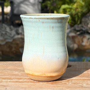 May include: A ceramic tumbler with a flared rim and a smooth, rounded body. The glaze features a gradient transition from a pale, speckled mint green at the top to a warm, soft peach hue at the base, with visible vertical drip patterns.