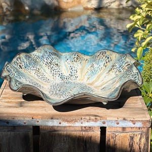 May include: A ceramic bowl with a wavy, ruffled rim and a textured surface featuring a mottled pattern of blue, cream, and dark brown tones. The interior displays an intricate, organic design with small, dark, elongated markings.