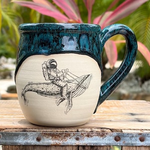 Peut inclure: Une tasse en céramique avec une glaçure brillante bleu sarcelle sur la partie supérieure et une base naturelle en crème mat. L'avant présente une illustration au trait noir d'un astronaute chevauchant une grande baleine avec une longe.