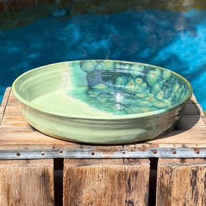 May include: A shallow, round ceramic bowl featuring a smooth, light green glaze on one side and a deep, crystalline blue-green glaze with circular, flower-like patterns on the other side, creating a distinct contrast in color and texture.