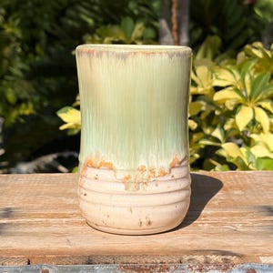 May include: A cylindrical ceramic tumbler featuring a smooth, pale green glaze on the upper portion that transitions into a textured, ribbed base with a light cream finish. The glaze exhibits a subtle drip effect where the green meets the cream.