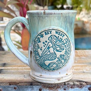 May include: A ceramic mug with a light blue and cream glaze. The mug features a circular emblem with a teal mermaid design and the words "KEY WEST". The handle is curved and the mug is on a wooden surface.