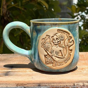 May include: A ceramic mug with a blue and green glazed finish featuring a circular tan medallion on the front. The medallion depicts a skeleton mermaid holding a cup with the text "Key West" and "Windy City" embossed on the surface.