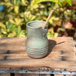 May include: A light green ceramic tumbler with a smooth, glossy glaze and subtle horizontal ridges near the base. The vessel features a small, integrated spout on the rim and contains a single, straight, light brown bamboo drinking straw.