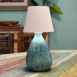 May include: A table lamp featuring a teardrop-shaped ceramic base with a glossy turquoise glaze and dark speckled accents. The lamp is topped with a white, conical, vertically pleated fabric lampshade that provides a clean, textured contrast.