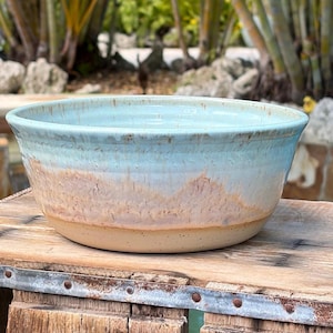 May include: A round ceramic bowl featuring a smooth, light blue glazed upper section that transitions into a textured, sandy-beige base. The glaze exhibits subtle vertical drips and variations in color, revealing the natural clay body.