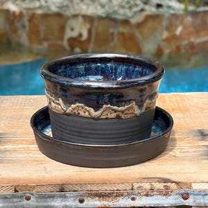 May include: A cylindrical ceramic planter with a matching saucer. The planter features a matte black base with horizontal ridges, a textured beige band in the middle, and a glossy, deep blue glazed rim that drips over the textured section.