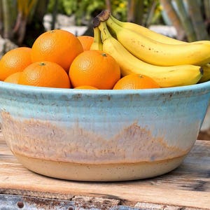 May include: A large, round ceramic bowl with a light blue glazed rim that transitions into a textured, earthy tan base. The bowl is filled with a cluster of bright yellow bananas and several round, vibrant orange citrus fruits.