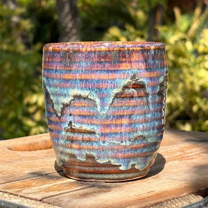 May include: A handmade ceramic tumbler with a unique, textured surface. The cup features horizontal stripes in shades of orange, blue, and purple, creating a vibrant, glazed finish. The tumbler is designed for beverages and is a decorative piece.
