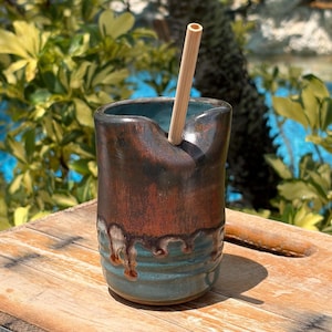 May include: A cylindrical ceramic tumbler with a notched rim, featuring a two-tone glaze of dark metallic brown on the upper portion and a teal blue base with a drip effect. A light-colored bamboo straw is inserted into the notched opening.