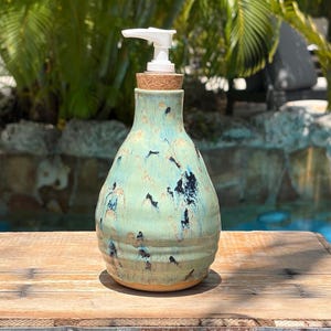 May include: A ceramic soap dispenser with a rounded, bulbous body featuring a light turquoise glaze with dark blue and tan speckled accents. The dispenser has a cork stopper at the neck and a white plastic pump mechanism at the top.