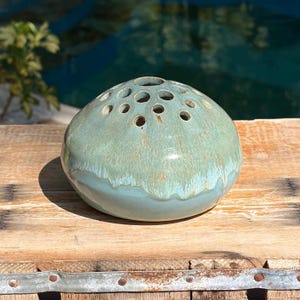 May include: A dome-shaped ceramic flower frog with a glossy finish in shades of sage green and light blue. The surface features multiple circular openings arranged across the top for stem placement, with a smooth, rounded base and drip glaze.