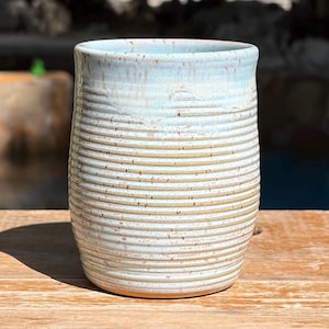 May include: A cylindrical ceramic tumbler featuring a light blue and cream glaze with a speckled finish. The exterior surface is textured with horizontal ridges that wrap around the entire body of the vessel, creating a tactile, ribbed appearance.