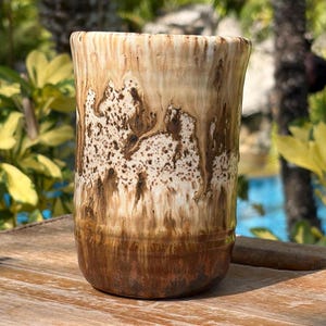 May include: A handcrafted ceramic tumbler with a unique, textured glaze. The colors transition from a dark brown base to a creamy beige at the top, with speckled and dripped patterns. The tumbler sits on a wooden surface.