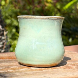 May include: A handmade ceramic mug with a rounded body and a slightly flared rim. The mug has a light green glaze with subtle blue undertones and a brown base. The rim has a darker, brownish-tan color.
